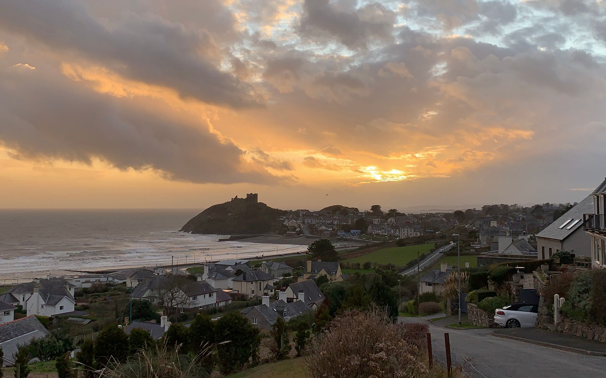 Vista da cidade costeira de Harlech, Gales, com o Castelo de Harlech em uma colina à beira-mar sob um céu alaranjado e nublado.
